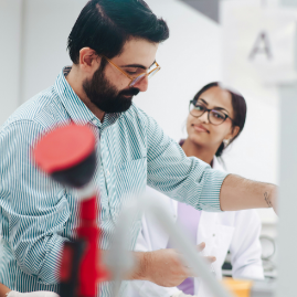 A male scientist in a green shirt and a female scientist in a lab coat working with equipment in a modern lab setting.