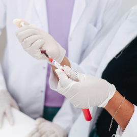 A close-up of two scientists in gloves using a syringe to carefully transfer a red liquid into a glass vial.