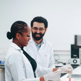 Two scientists in white lab coats and gloves smiling while collaborating on an experiment in a bright laboratory.